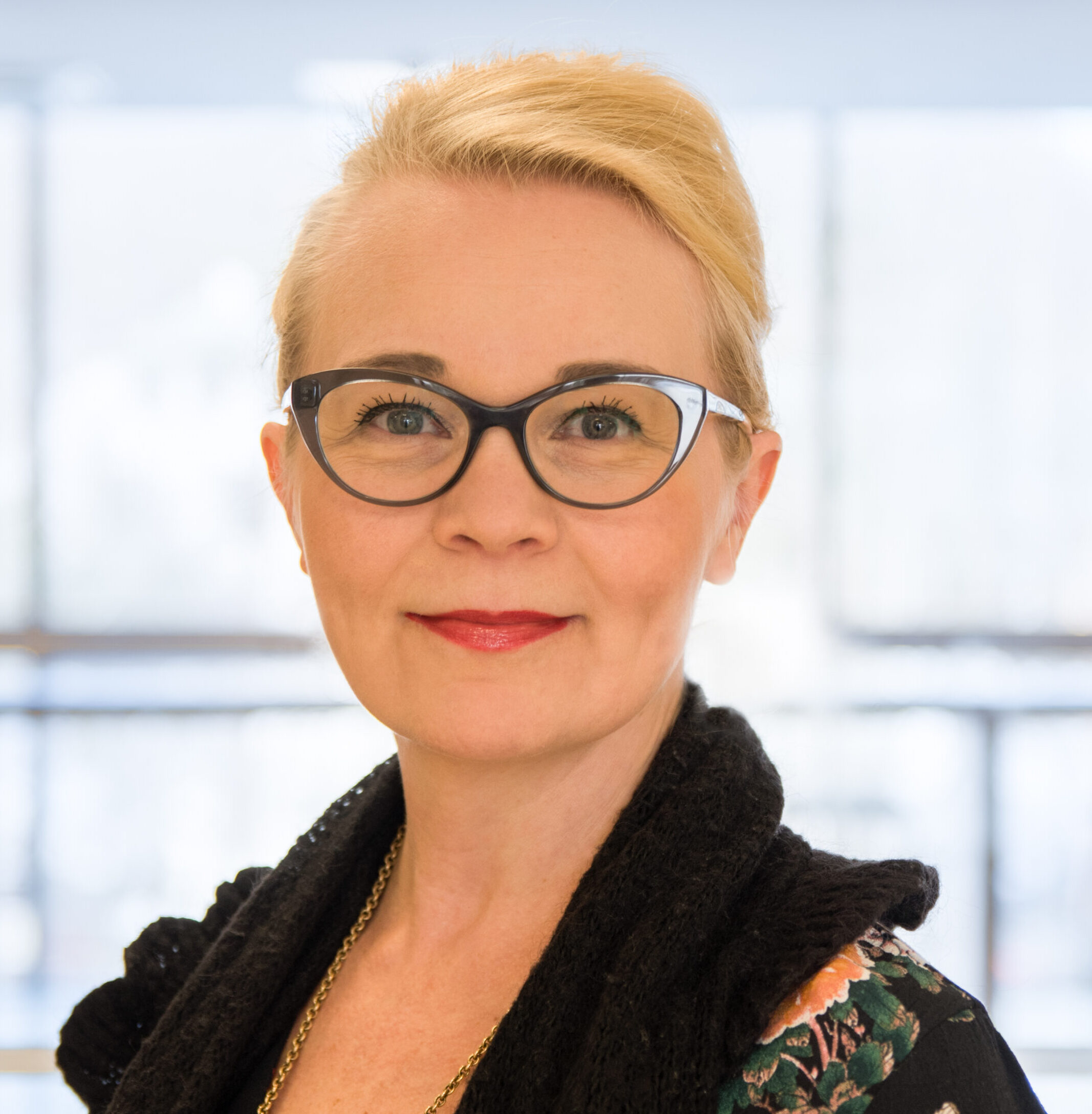 Kasvokuvassa hillitysti hymyilevä vaaleatukkainen, tummaan asuun pukeutunut nainen, jolla tummat silmälasit ja hiukset sidottu kiinni.