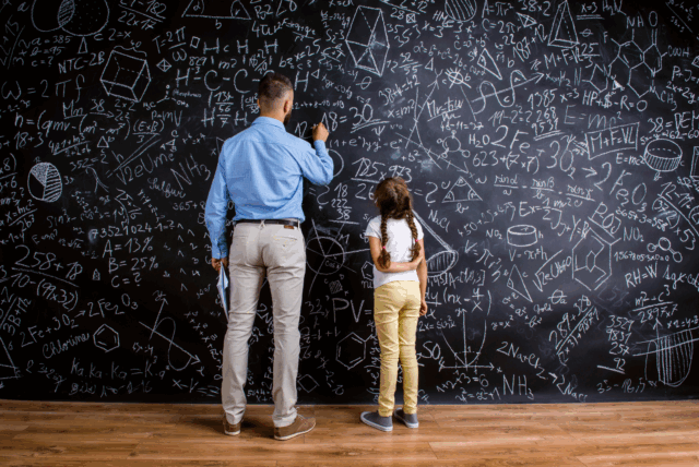 Kuvassa selin täyteen piirretyllä liitutaululla vaaleisiin housuihin ja vaaleansiniseen paitaan pukeutunut mies ja vaaleisiin housihin ja valkoiseen t-paitaan pukeutunut tummalettinen pikkutyttö.