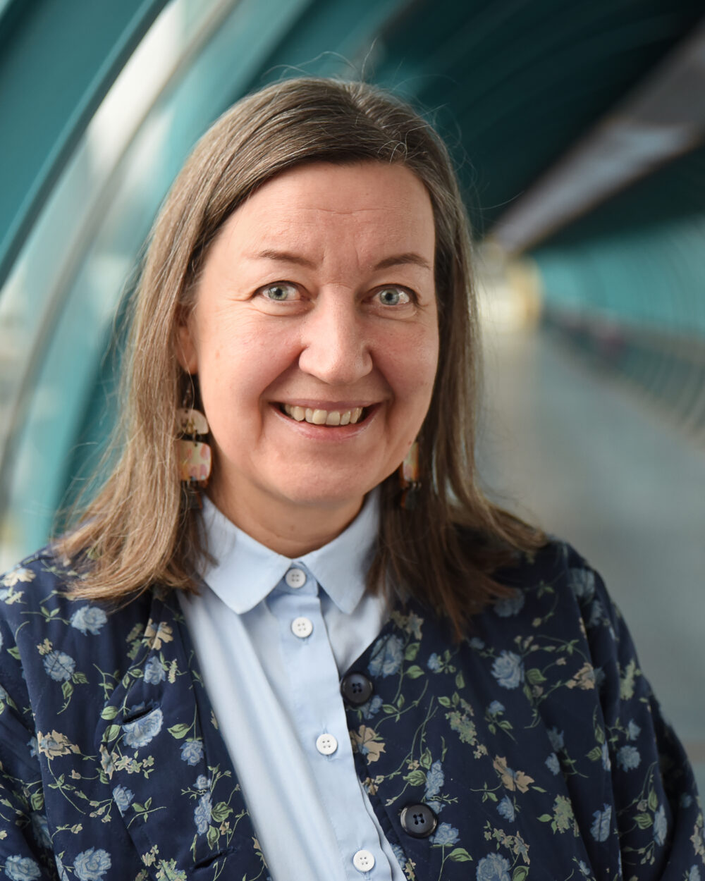 Hymyilevä ruskeatukkainen naisoletettu putkimaisessa ikkunallisessa käytävässä pukeutuneena vaaleansiniseen kauluspaitaan ja tummansiniseen kirjavaan jakkuun.