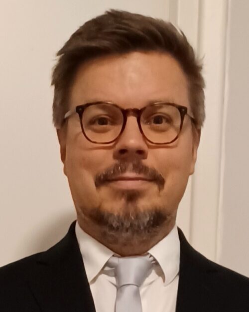 Tiukasti rajatussa kasvokuvassa muovisankaisiin silmälaseihin sonnustautunut nuorehko miesoletettu, jolla valkoinen kauluspaita ja harmaa solmio.